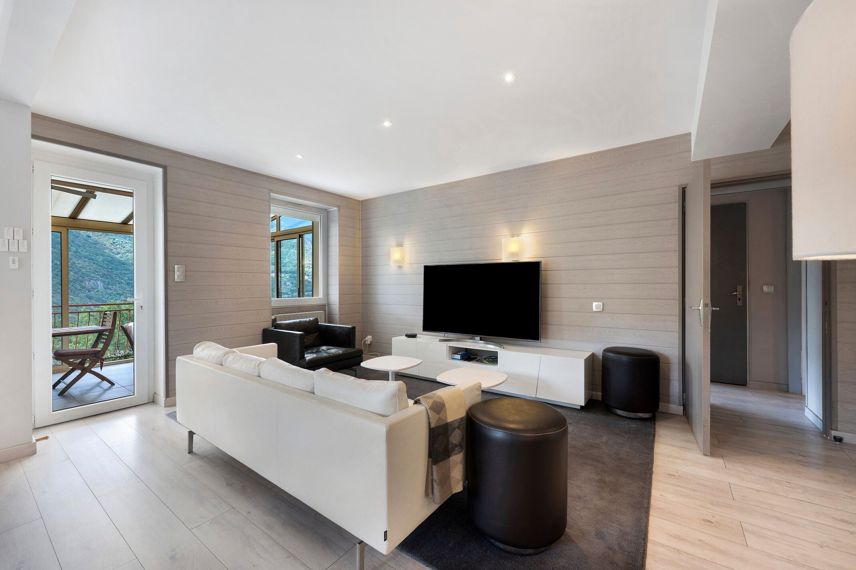 En moderne stue med sofa, sofabord, et fladskærms-tv og en balkon med udsigt til bjergene.
