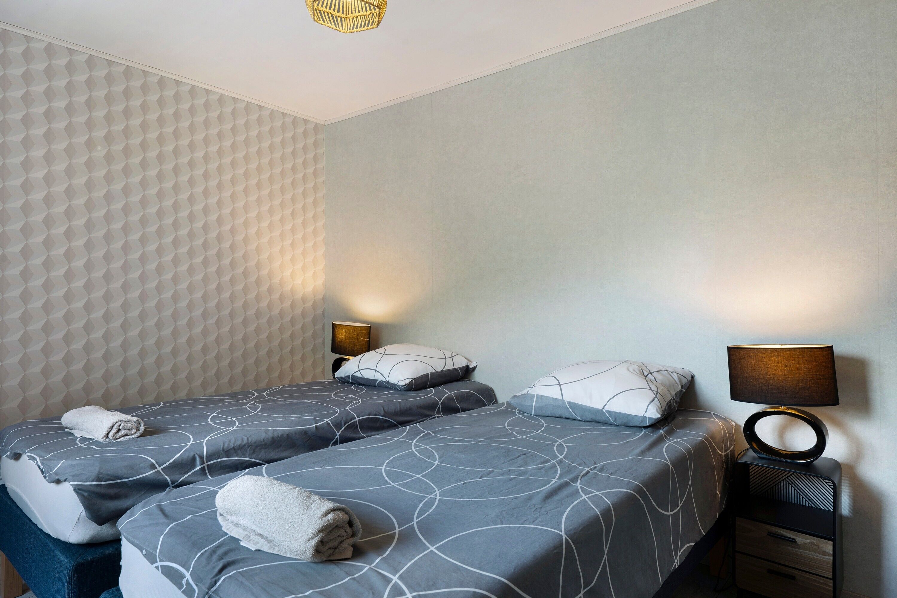 Ein Hotelzimmer mit zwei Betten, jeweils mit grauer Bettdecke und weißen Kissen, einem Nachttisch mit Lampe sowie einer strukturierten Wand.