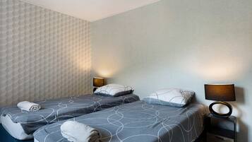Ein Hotelzimmer mit zwei Betten, jeweils mit grauer Bettdecke und weißen Kissen, einem Nachttisch mit Lampe sowie einer strukturierten Wand.