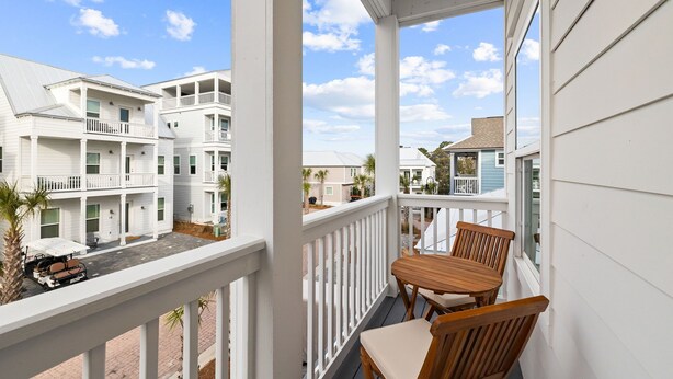 A balcony with wooden chairs and a table, overlooking a street with multi-story buildings. - Dune Allen/30A! Walk to Beach/Coffee/Dinner! Bikes! (Santa Rosa Beach)