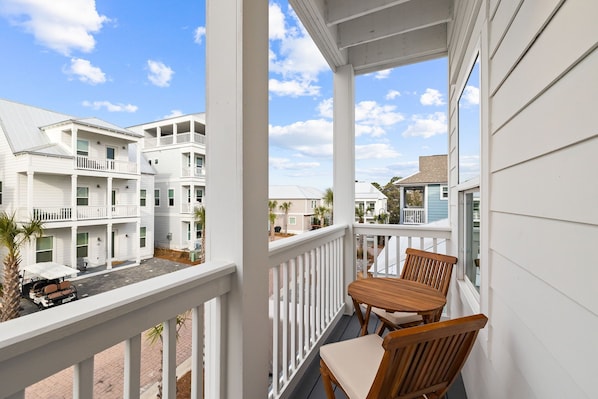 A balcony with wooden chairs and a table, overlooking a street with multi-story buildings. - Dune Allen/30A! Walk to Beach/Coffee/Dinner! Bikes! (Santa Rosa Beach)
