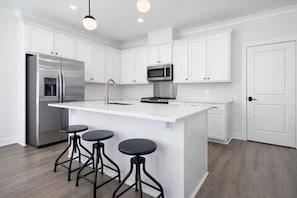 A modern kitchen with white cabinetry, a stainless steel refrigerator, a microwave, a sink, and a bar area with stools.