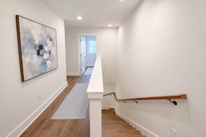 A hallway with a wooden floor, a white wall, and a staircase with a wooden handrail.