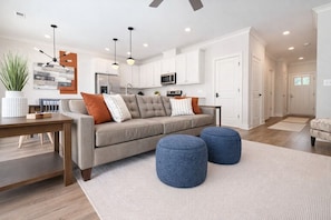 Een moderne woonkamer met een bank, poufs, een salontafel en een keuken op de achtergrond.