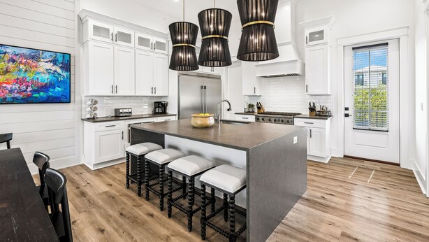 A modern kitchen with white cabinetry, a large island with bar stools, pendant lighting, and a view of greenery outside.