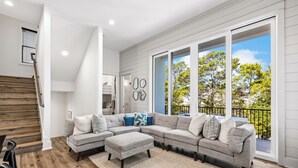 A modern living room with a sectional sofa, a wooden staircase, and large windows with a view of trees.