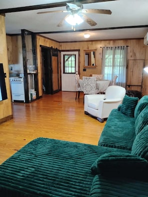 Un salon confortable avec un canapé vert, une table à manger avec des chaises, un coin cuisine équipé d’une cuisinière et d’un réfrigérateur, et un ventilateur de plafond.