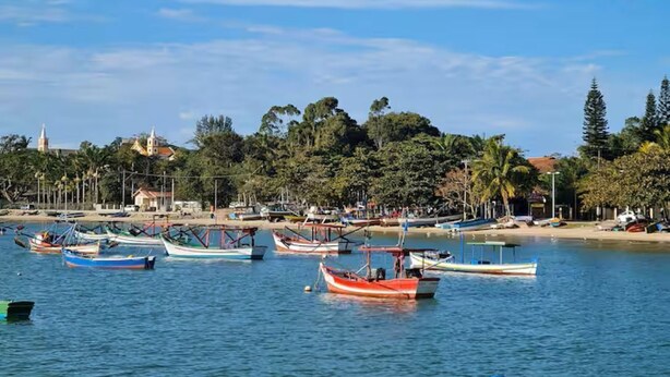 A coastal scene with boats docked near a beach, trees, and buildings in the background. - Family apartment, calm and welcoming, with beach and park nearby. (Penha SC)