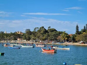 Escena costera con barcos amarrados cerca de una playa, árboles y edificios al fondo.