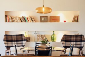 A dining area with a round table, two chairs with plaid upholstery, a hanging pendant light, and a shelf above with books and a framed picture.