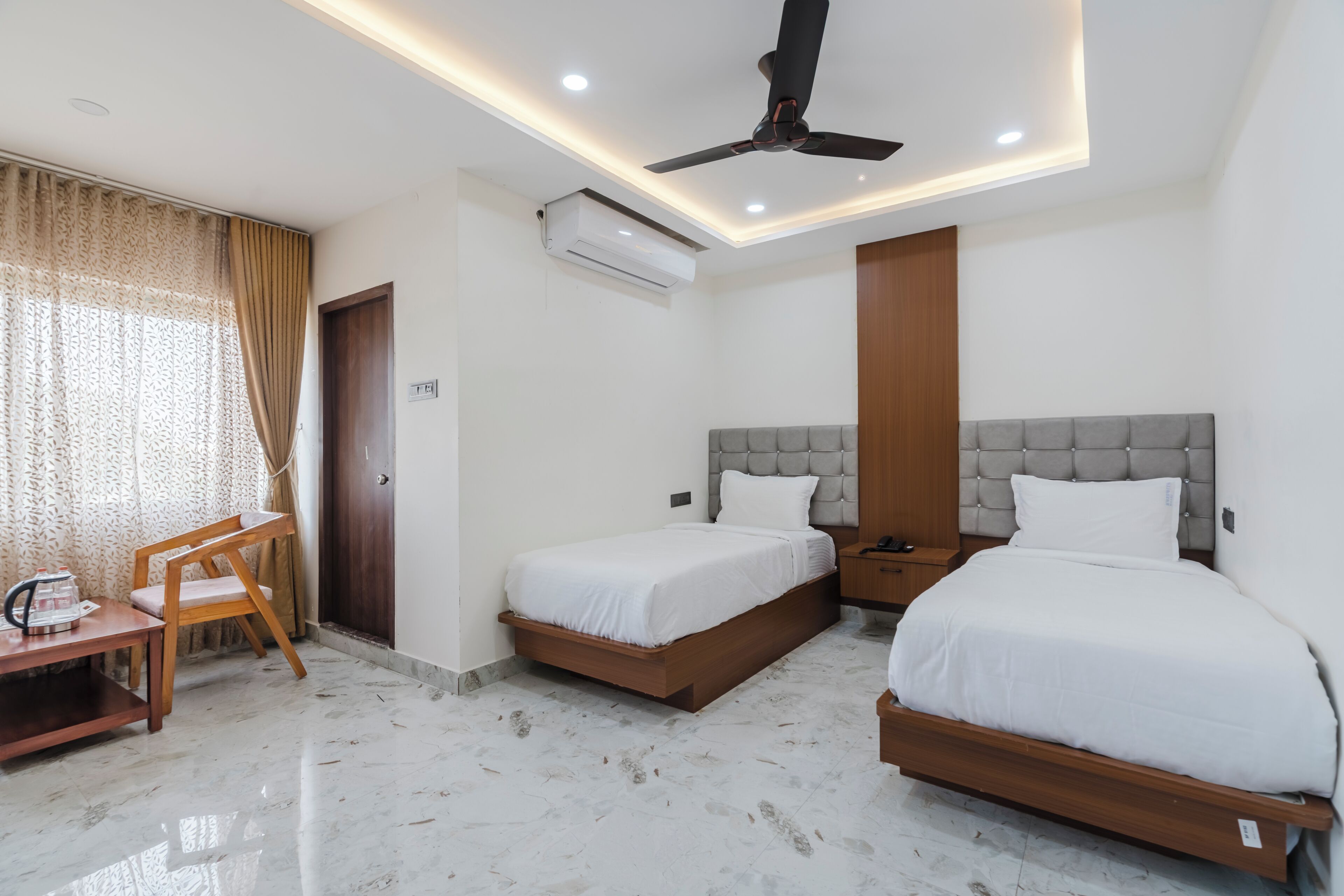 A hotel room with two beds, a ceiling fan, an air conditioning unit, a wooden nightstand, a desk with a chair, and a window with curtains.