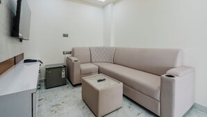 A modern living room with a beige sectional sofa, a glass-top coffee table, a small refrigerator, and a flat-screen TV mounted on the wall. - Hotel Rasaballa grand (Kothagudem)