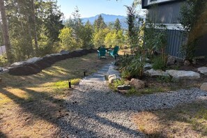 A backyard with a gravel path, two green chairs, a wooden deck, and a stone wall.