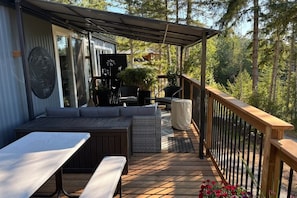 Une terrasse dotée d’un canapé, de chaises et d’une table, entourée d’une rambarde en bois et offrant une vue sur une forêt.