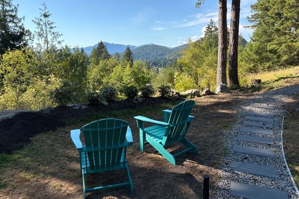Two green Adirondack chairs on a gravel path with a view of a forest and mountains in the background.