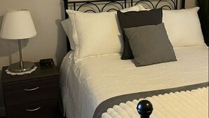 Ein ordentlich bezogenes Bett mit weißer Decke und Kissen, einem schwarzen Metall-Kopfteil, einem Nachttisch mit Lampe und einem gemusterten Teppich.