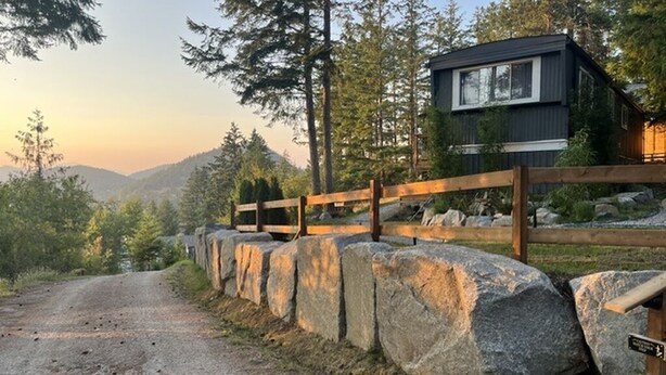 Eine Holzhütte mit Holzzaun und großen Steinen davor, umgeben von Bäumen und Bergen.