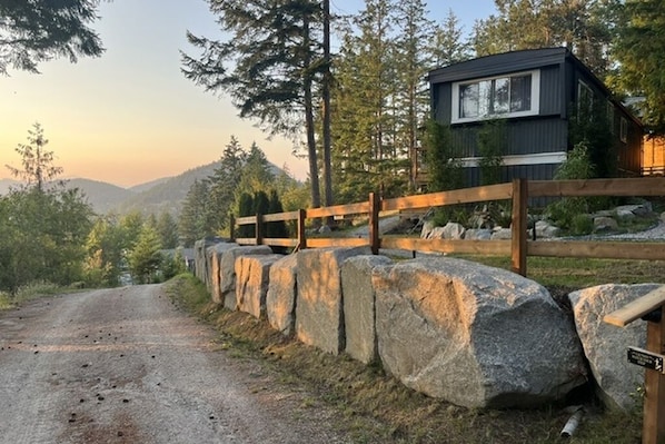 A cabin with a wooden fence and large rocks in front, surrounded by trees and mountains.
