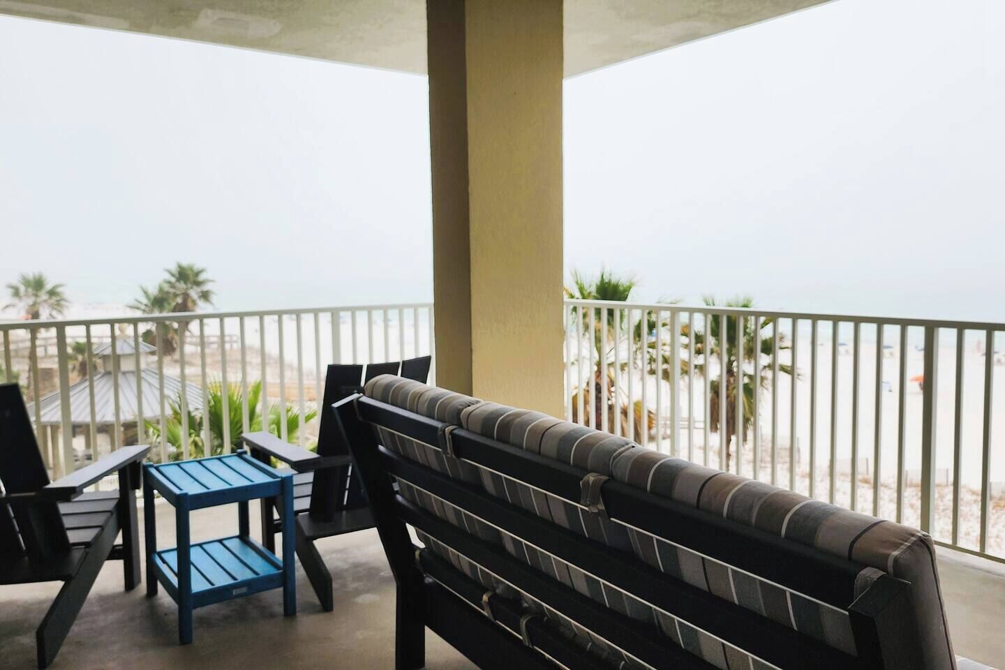 A balcony with a sofa, a small table, and chairs, overlooking a beach with palm trees.