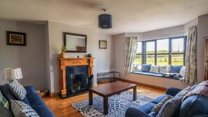 A living room with a fireplace, blue sofas, a wooden coffee table, and a large window with a view of the outdoors. - Strand House (Bannow)