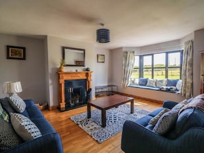 A living room with a fireplace, blue sofas, a wooden coffee table, and a large window with a view of the outdoors. - Strand House (Bannow)