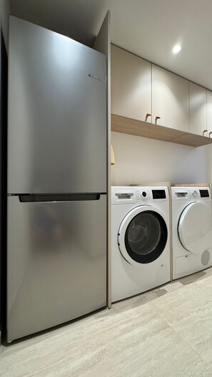 Une cuisine équipée d’un réfrigérateur en acier inoxydable, d’un lave-linge et d’une étagère en bois située au-dessus des appareils.