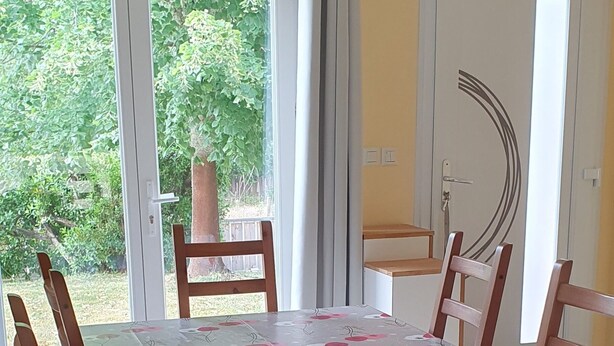Ruokailutila, jossa on neljälle katettu pöytä, ja jonka ympärillä on puisia tuoleja, kattovalaisin sekä lasiovesta avautuva näkymä ulos.