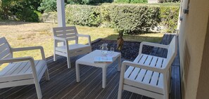 A covered outdoor seating area with white plastic chairs and a table, surrounded by a wooden deck and a hedge. - House with a garden (fenced), in a very quiet location, overlooking the forest (Lacanau)