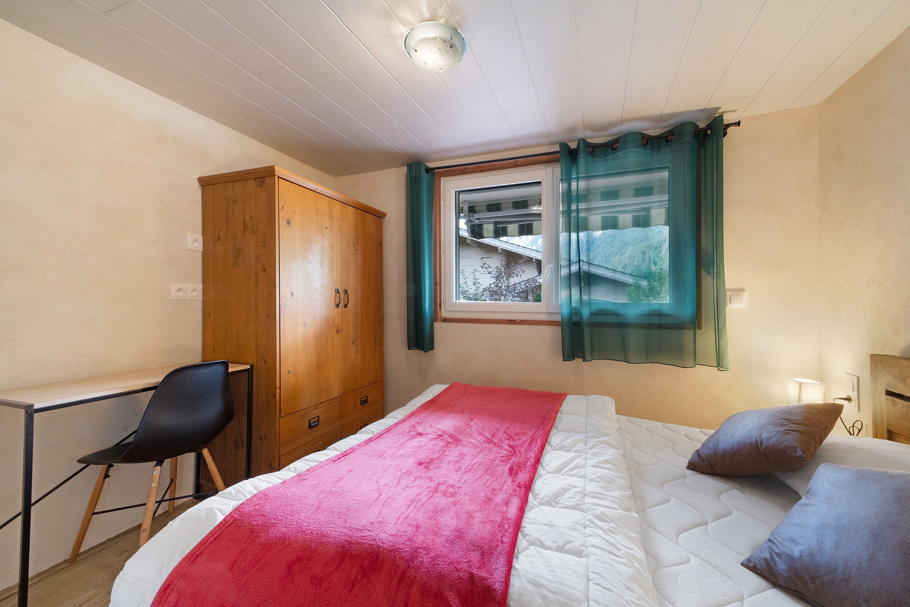 A bedroom with a bed, a desk, a chair, a wardrobe, and a window with curtains.