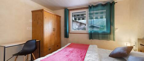 A bedroom with a bed, a desk, a chair, a wardrobe, and a window with curtains.