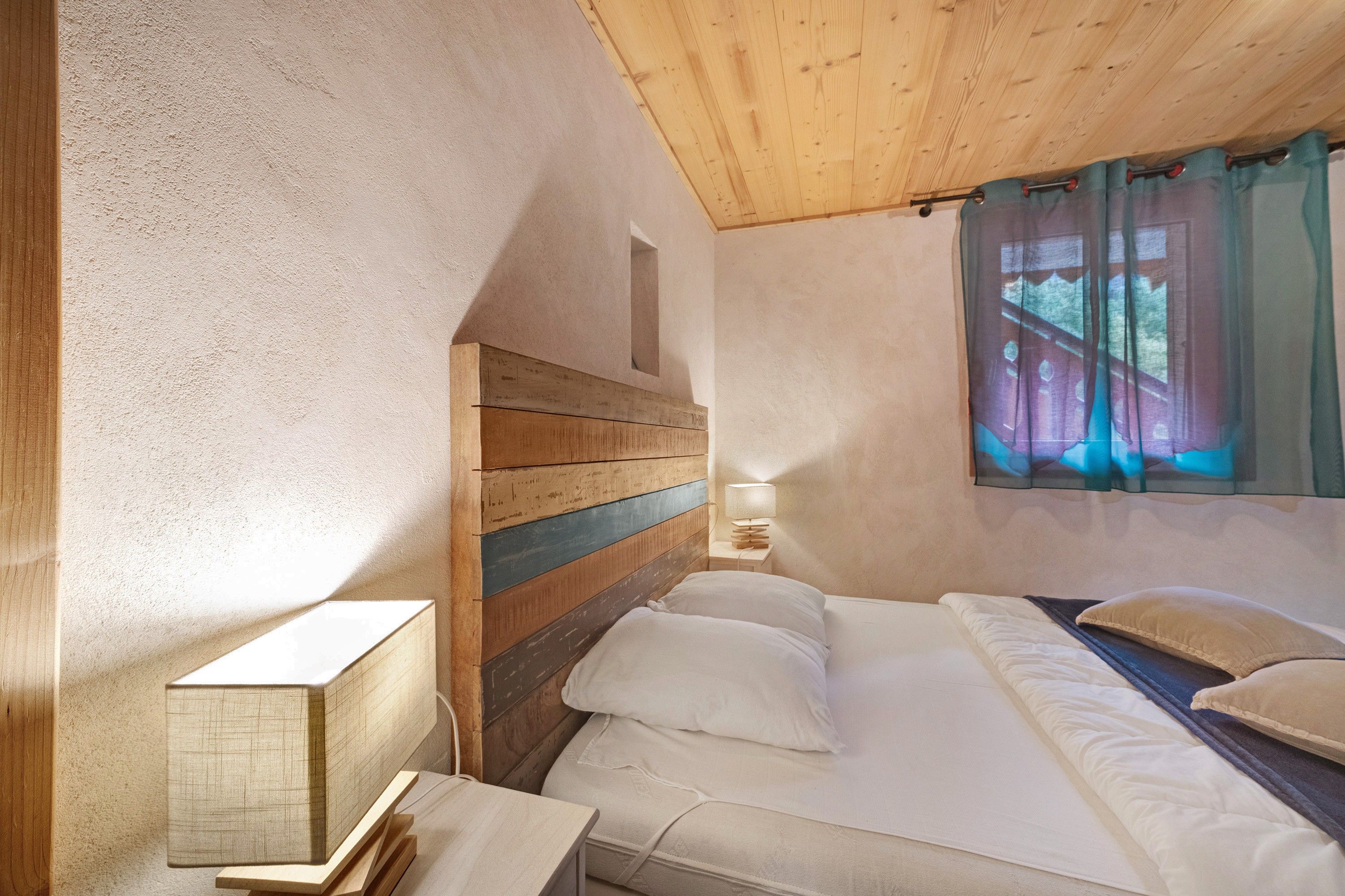 A bedroom with a wooden headboard, a bedside table with a lamp, and a window with curtains.