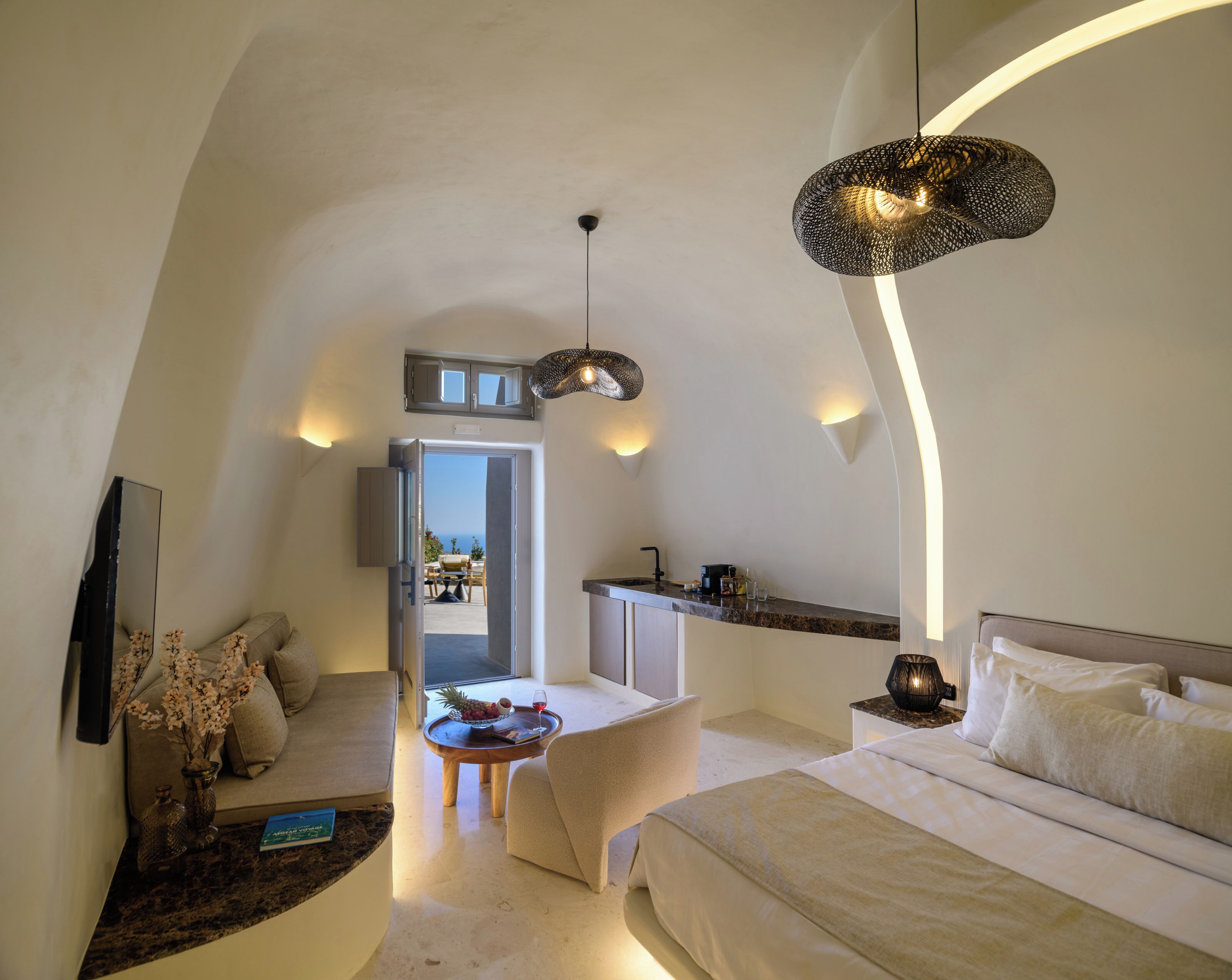 Un dormitorio moderno y bien iluminado con una cama grande, una zona de estar con una mesa, una cocina con mostrador y vistas a través de una puerta abierta.