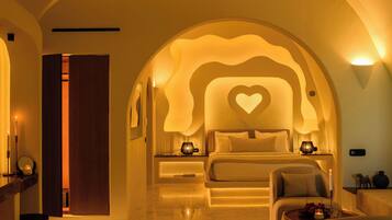 A hotel room with a bed, a chair, a table, and a decorative heart-shaped light on the wall.
