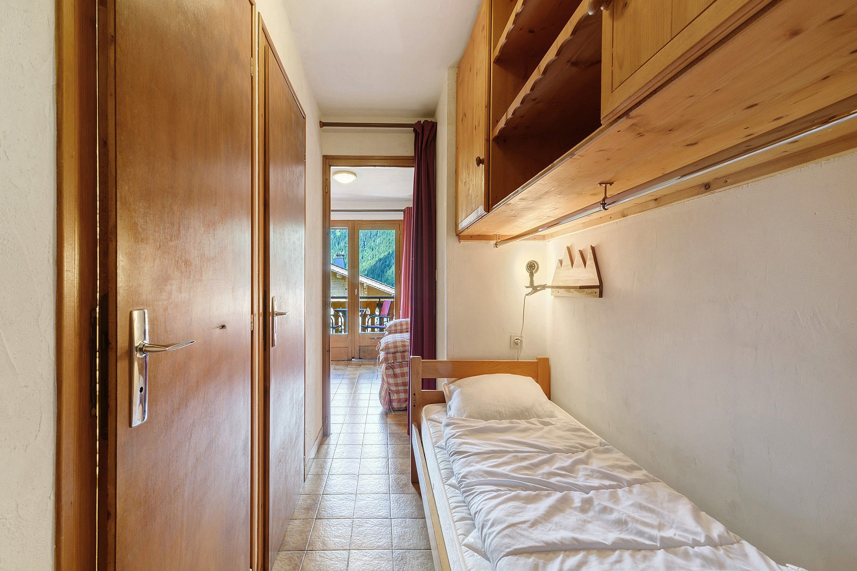 Ein schmaler Flur führt zu einem Schlafzimmer mit einem Bett, einem Holzschrank und einem Fenster mit Vorhängen.