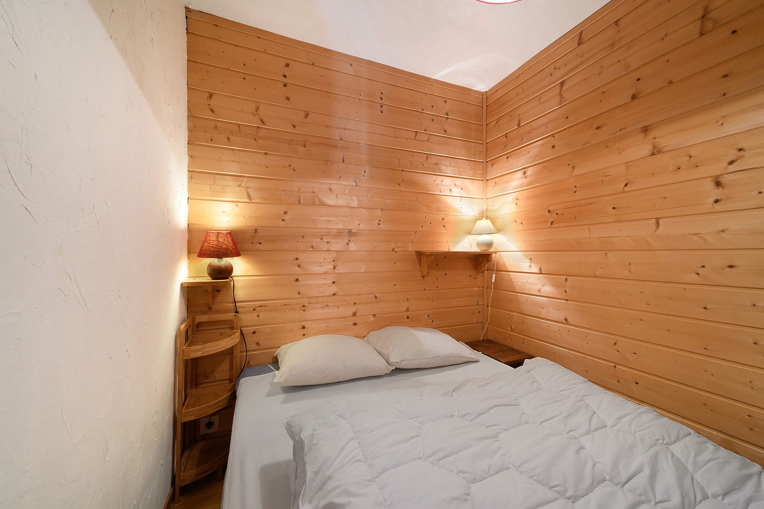 A small, cozy room with wooden walls, a bed with white bedding, a nightstand with a lamp, and a wall-mounted shelf.