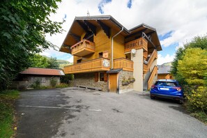 A two-story wooden building with balconies, a stone wall, and a parked car. - 2-Room Apartment for 4 – Châtel – Balcony, Parking, Near the Slopes (Châtel)