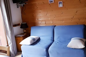 A room with a blue sofa, two pillows, a lamp, a TV, and wooden walls. - Apartment for 5 in Châtel – Near Slopes, Balcony, Free Private Parking (Châtel)