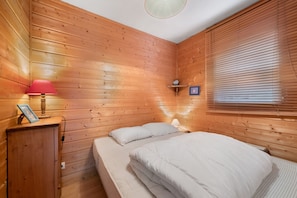 A wooden-paneled room with a bed, a nightstand, a lamp, and a window with blinds. - Apartment for 5 in Châtel – Near Slopes, Balcony, Free Private Parking (Châtel)
