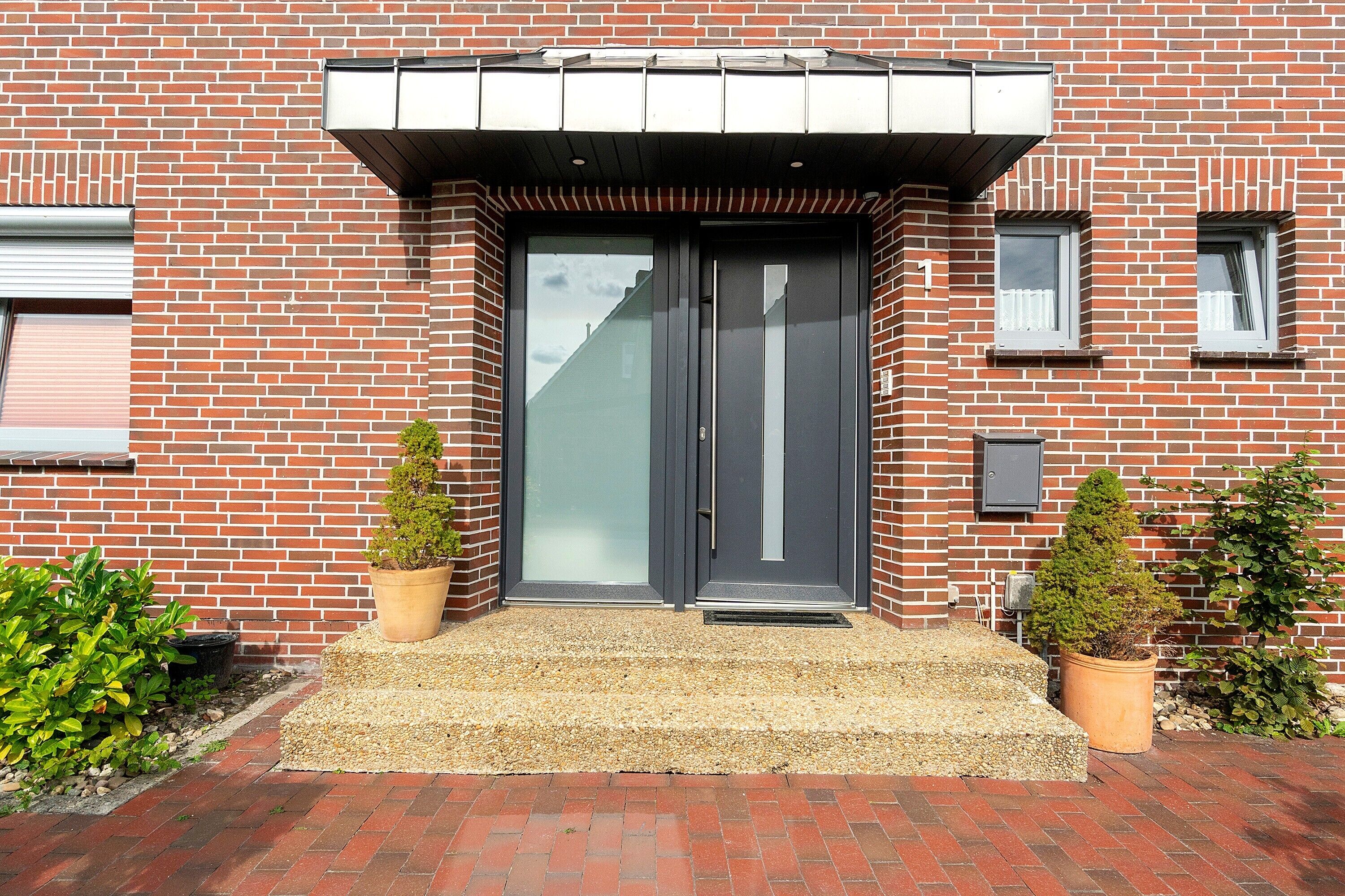 A modern brick building with a dark entrance door, a small window, and two potted plants on the ground.