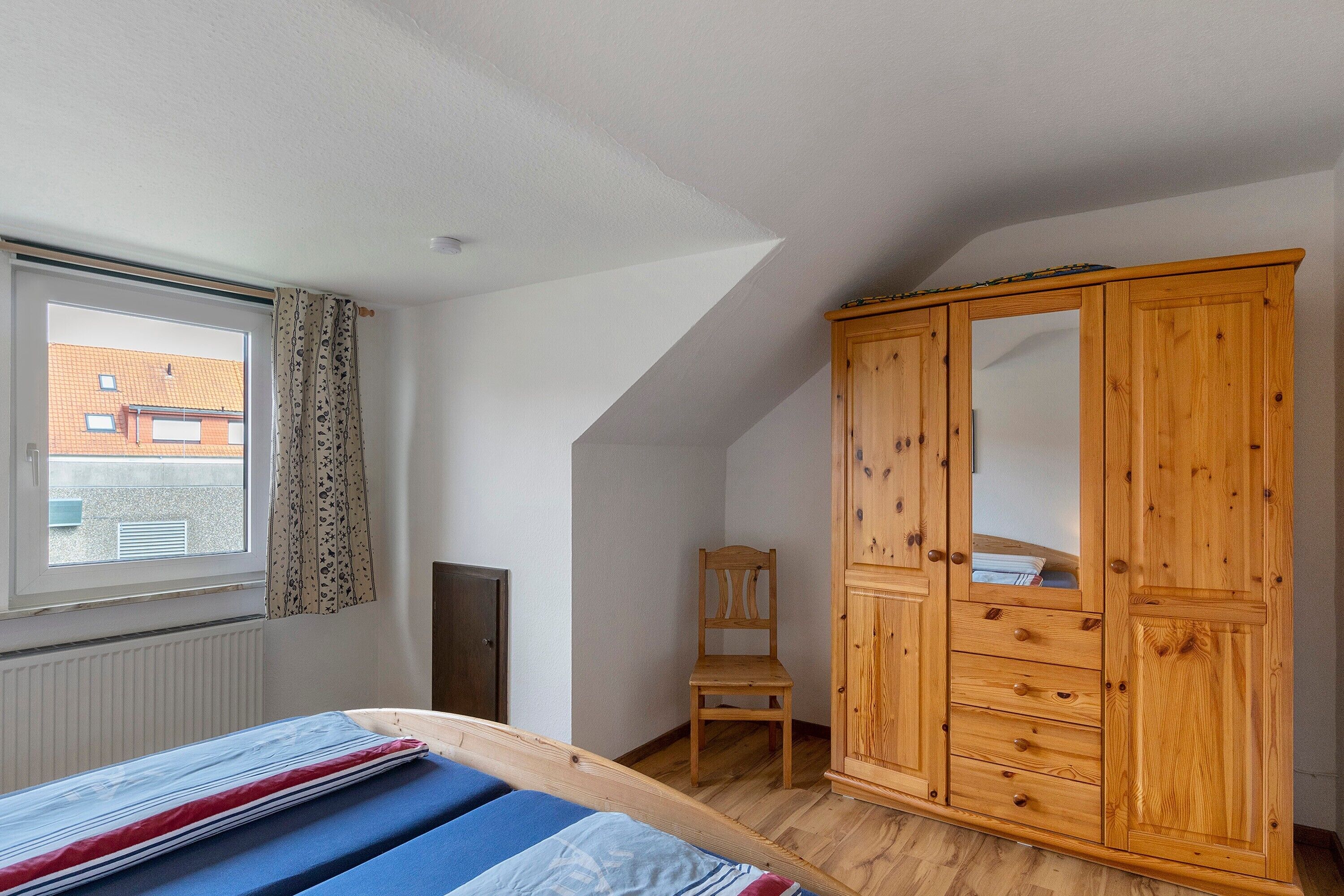Ein Schlafzimmer mit schrägem Dach, einem Holzschrank, einem Einzelbett mit gestreifter Matratze, einem Sessel und einem Fenster mit Vorhängen.