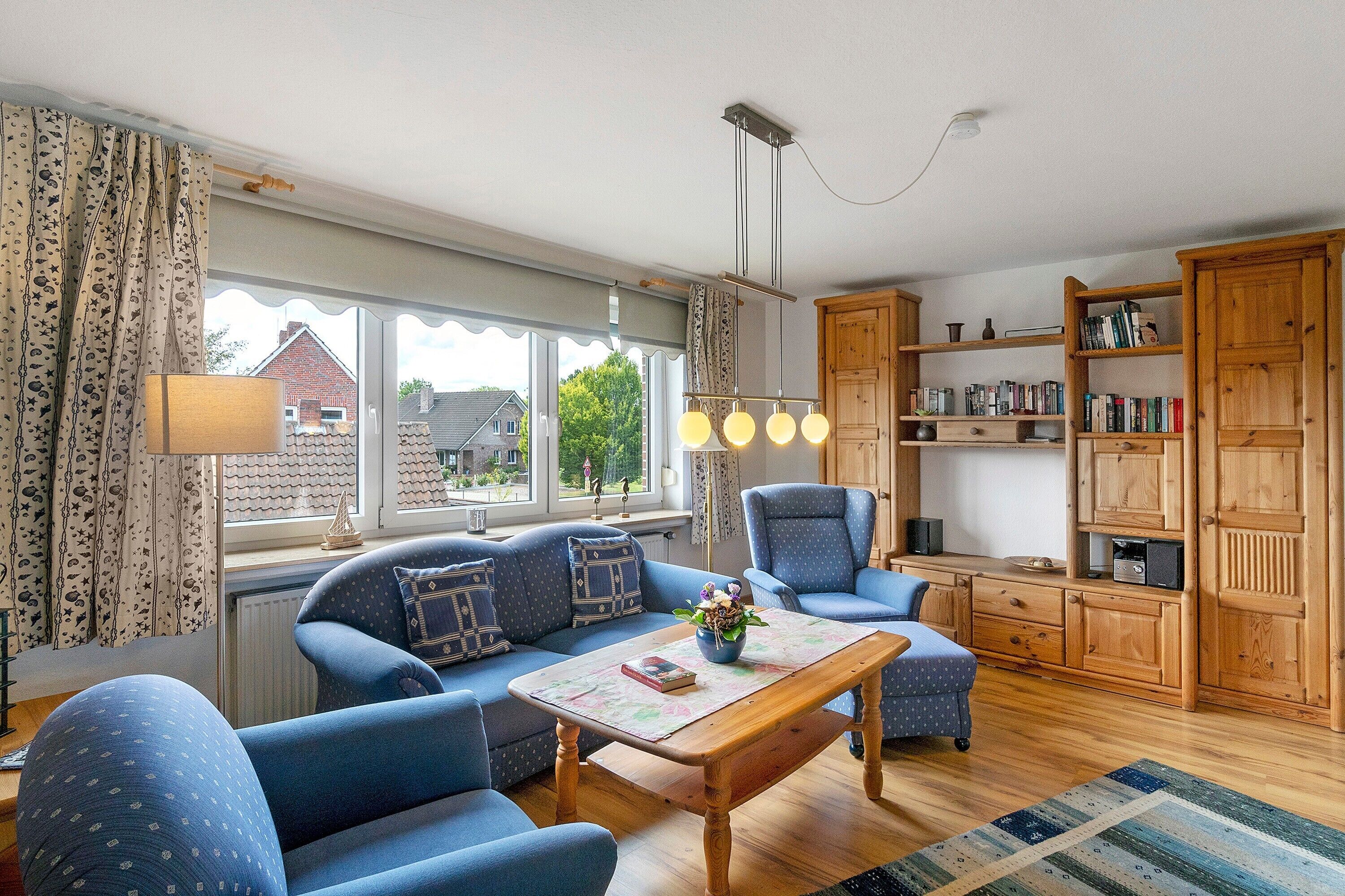 Ein gemütliches Wohnzimmer mit einer Couch, einem Sessel und einem Holztisch. Ein Bücherregal voller Bücher und ein Fenster mit Vorhängen sind ebenfalls vorhanden.