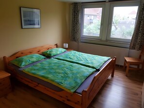 Ein Zimmer mit einem hölzernen Bett, einem Nachttisch, einem Stuhl, einem Vorhangfenster und einer Heizung.