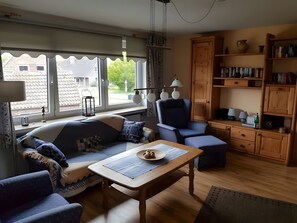 Ein gemütliches Wohnzimmer mit Sofa, Sessel und Tisch, großen Fenstern mit Aussicht sowie einem Bücherregal aus Holz.