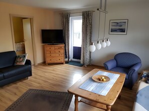Een woonkamer met een bank, een tafel met een tafelonderzetter, een televisie op een houten kast en een stoel.