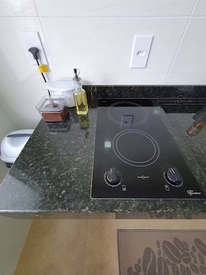 Een keuken met een zwarte inductiekookplaat, een witte stopcontact en een fles olie, plus een bak met eten.
