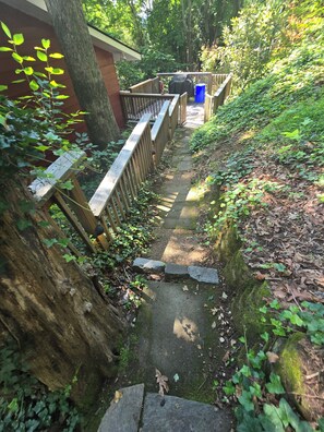 Ein schmaler Steinweg, gesäumt von hölzernen Geländern, der durch ein bewaldetes Gebiet führt, mit einem Gebäude im Hintergrund.