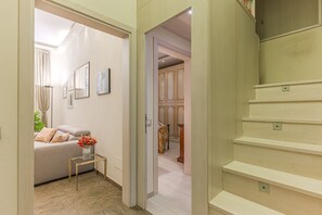 A hallway with a staircase, a sofa with cushions, a small table with a vase, and framed pictures on the wall.