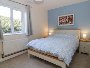 Ein Schlafzimmer mit einem Bett, Nachttischen, einem Heizkörper, einem Fenster mit Vorhängen und einem Blumenbild an der Wand.