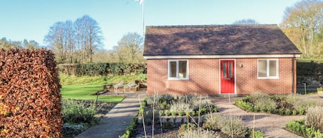 A red-brick house with a red door, a white window, and a garden with various plants and a pathway.