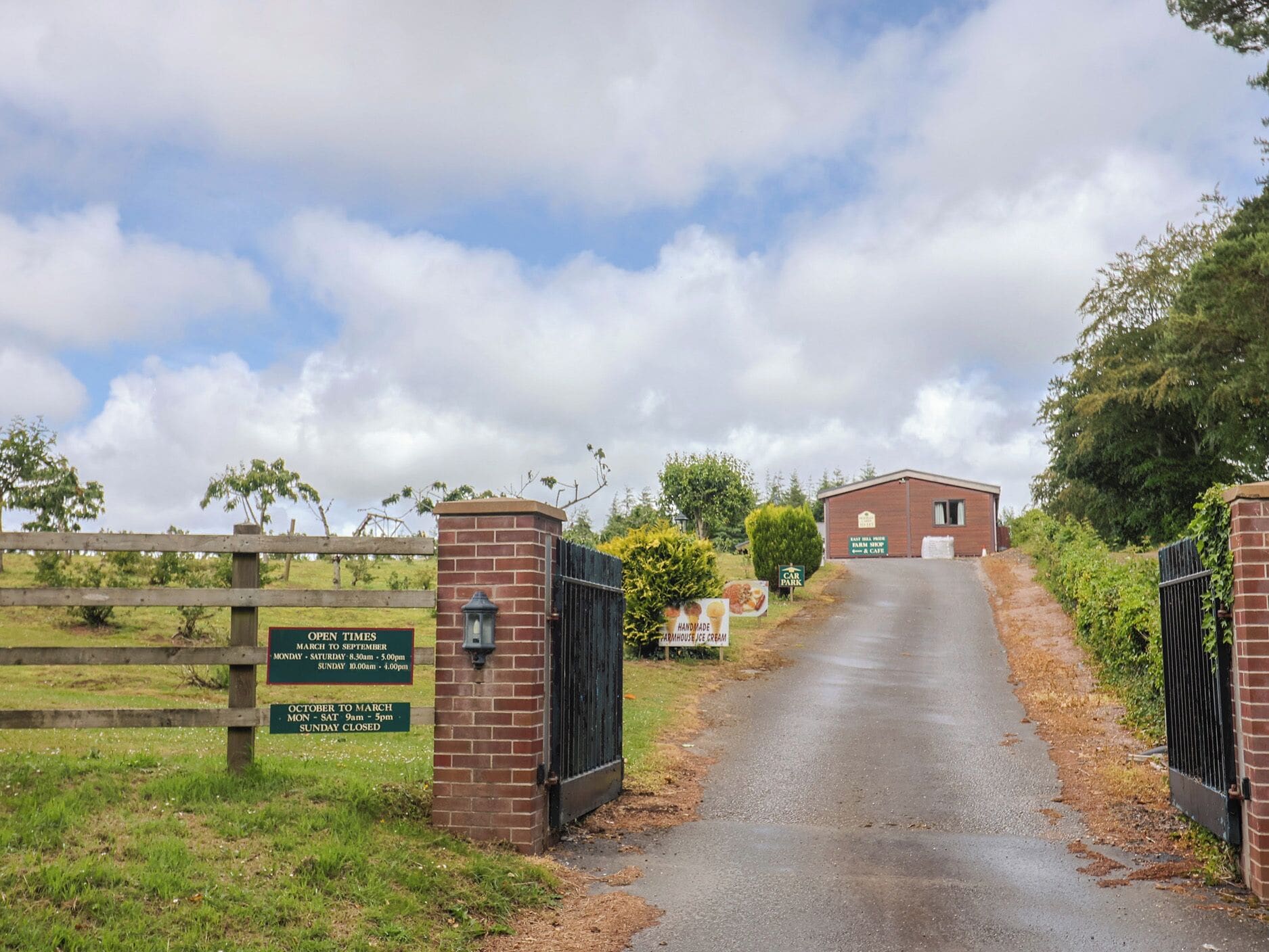 Una entrada rural con una cancela, un cartel que dice 'Car Park' y un edificio con el letrero 'Open Times'.